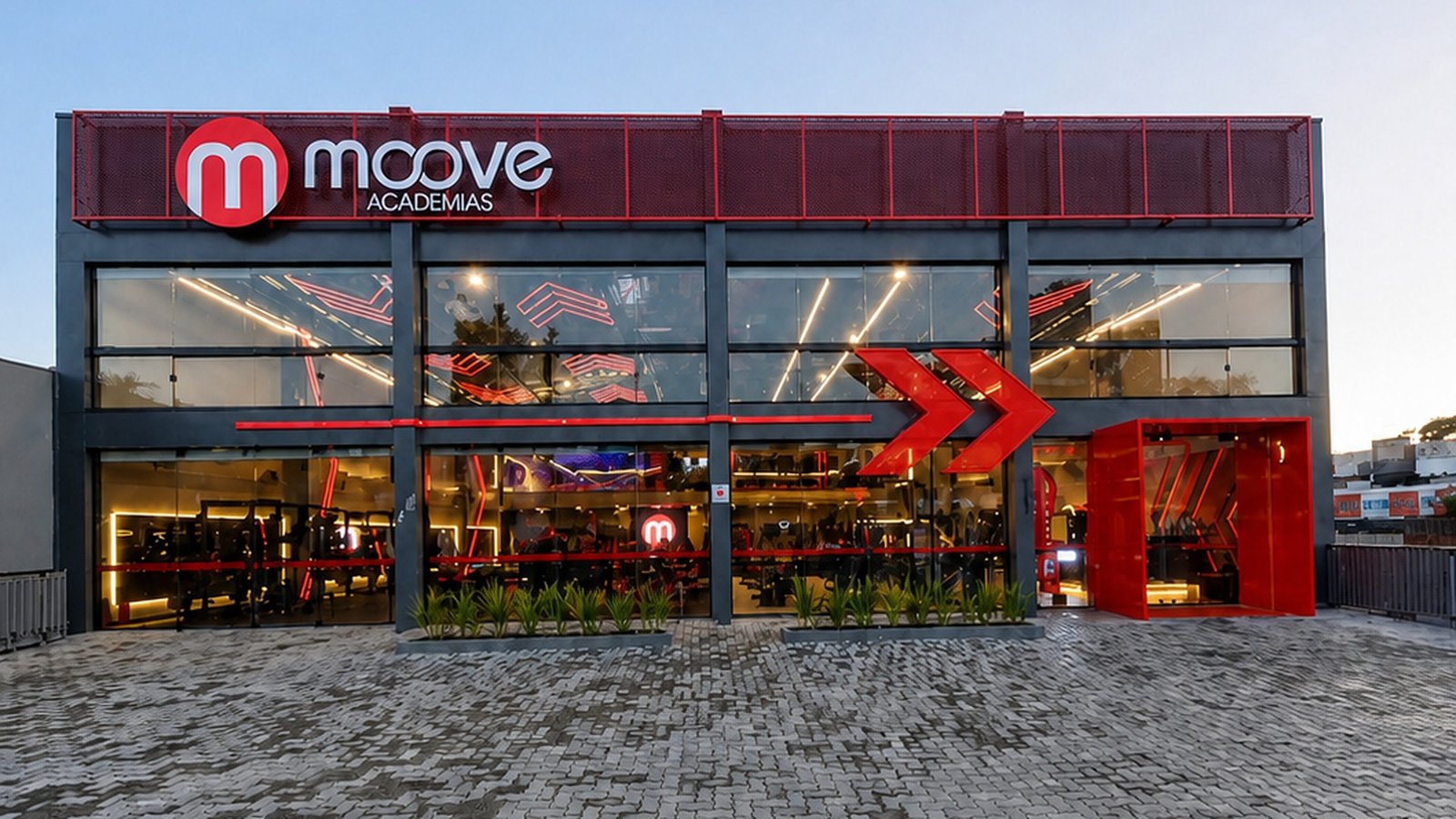 Fachada da Moove Academias Montenegro, com estrutura envidraçada e iluminação interna visível.